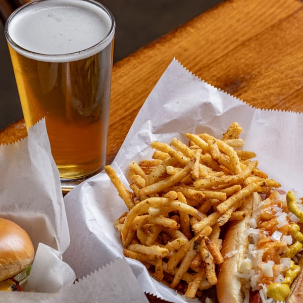 A hot dog with onions and mustard, seasoned fries, and a glass of beer on a wooden table.