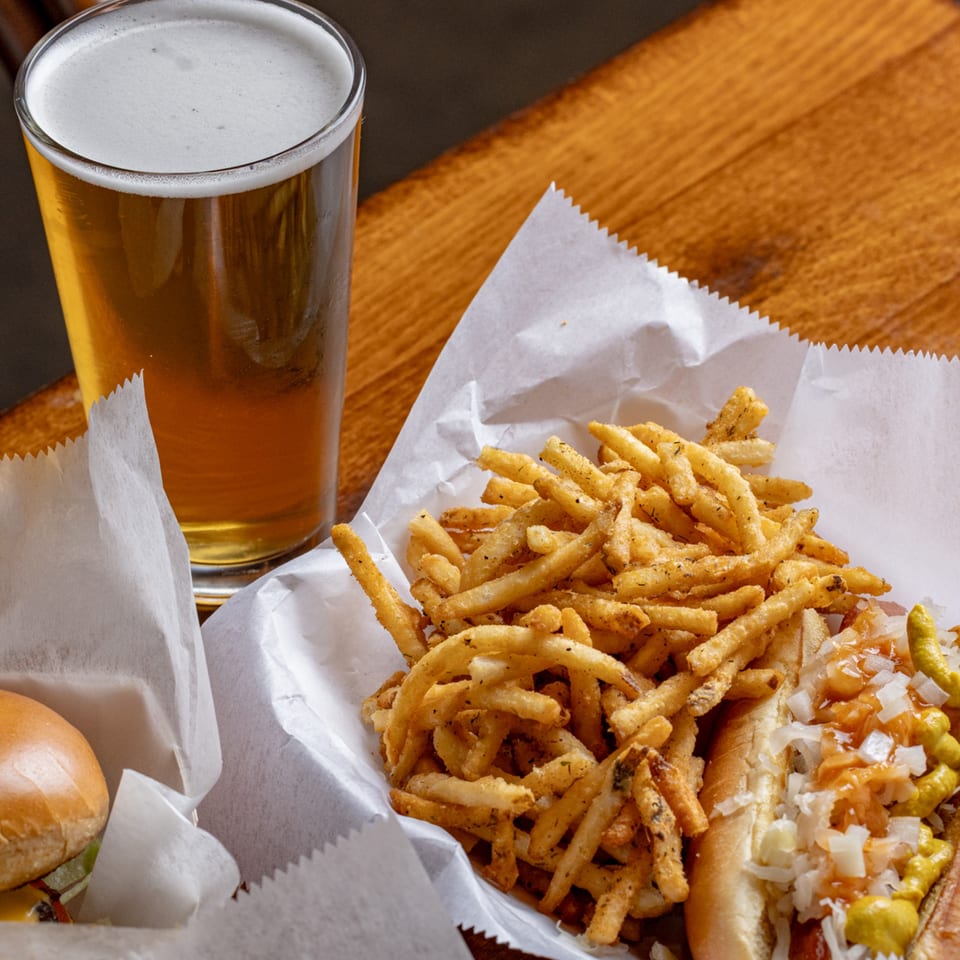 A hot dog with onions and mustard, seasoned fries, and a glass of beer on a wooden table.