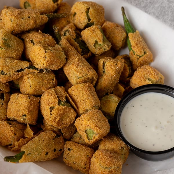 Crispy fried okra pieces with a side of creamy ranch dipping sauce in a white paper-lined basket.