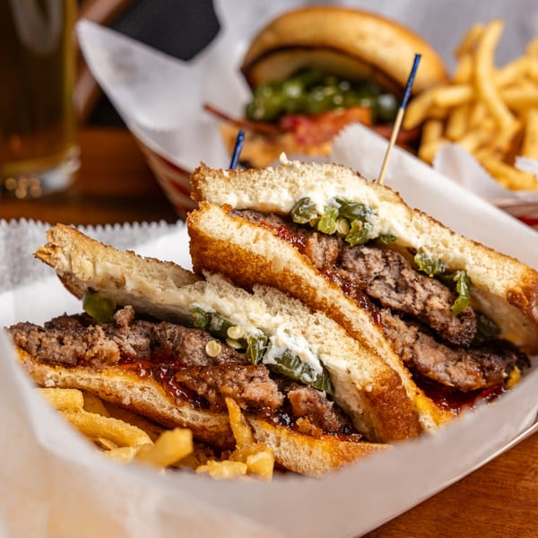 A grilled cheese burger with jalapeños and sauce, served with fries in a paper-lined basket.